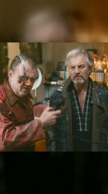 Two men in a cluttered room, one with a robotic glass eye holding a mask